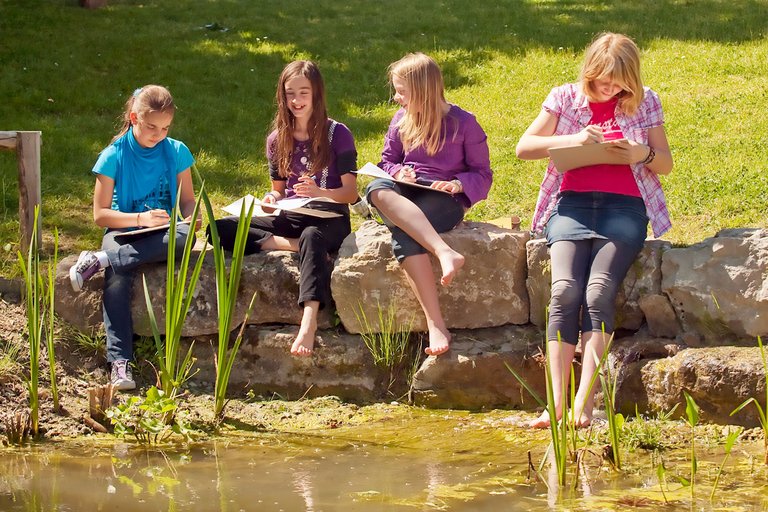 Students sit on a wall at the edge of a pond and paint.
