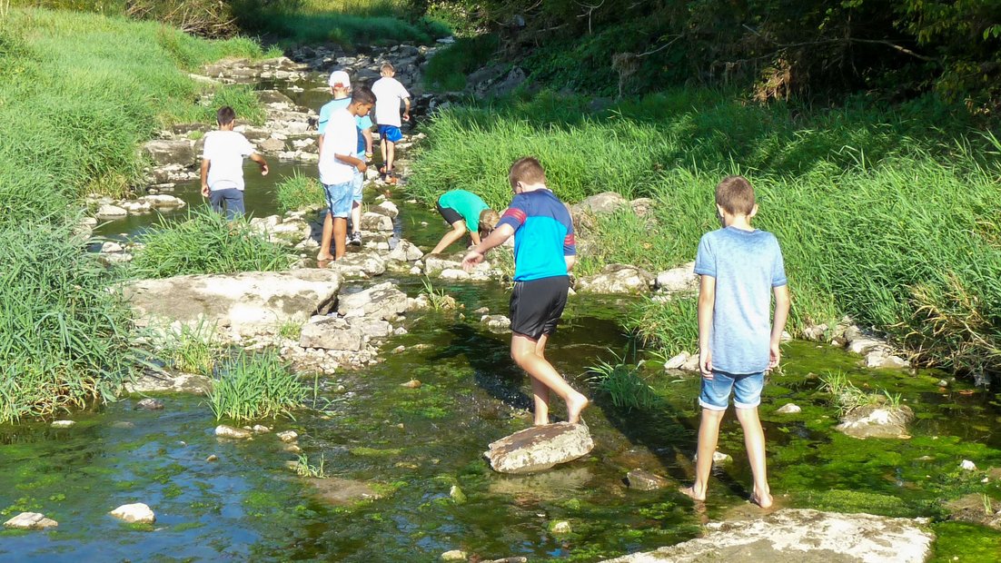 Younger students explore a shallow riverbed with large stones during a day of hiking.
