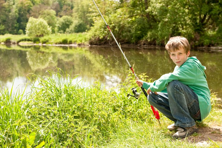 Boy sits on the river bank with a fishing rod in his hand.